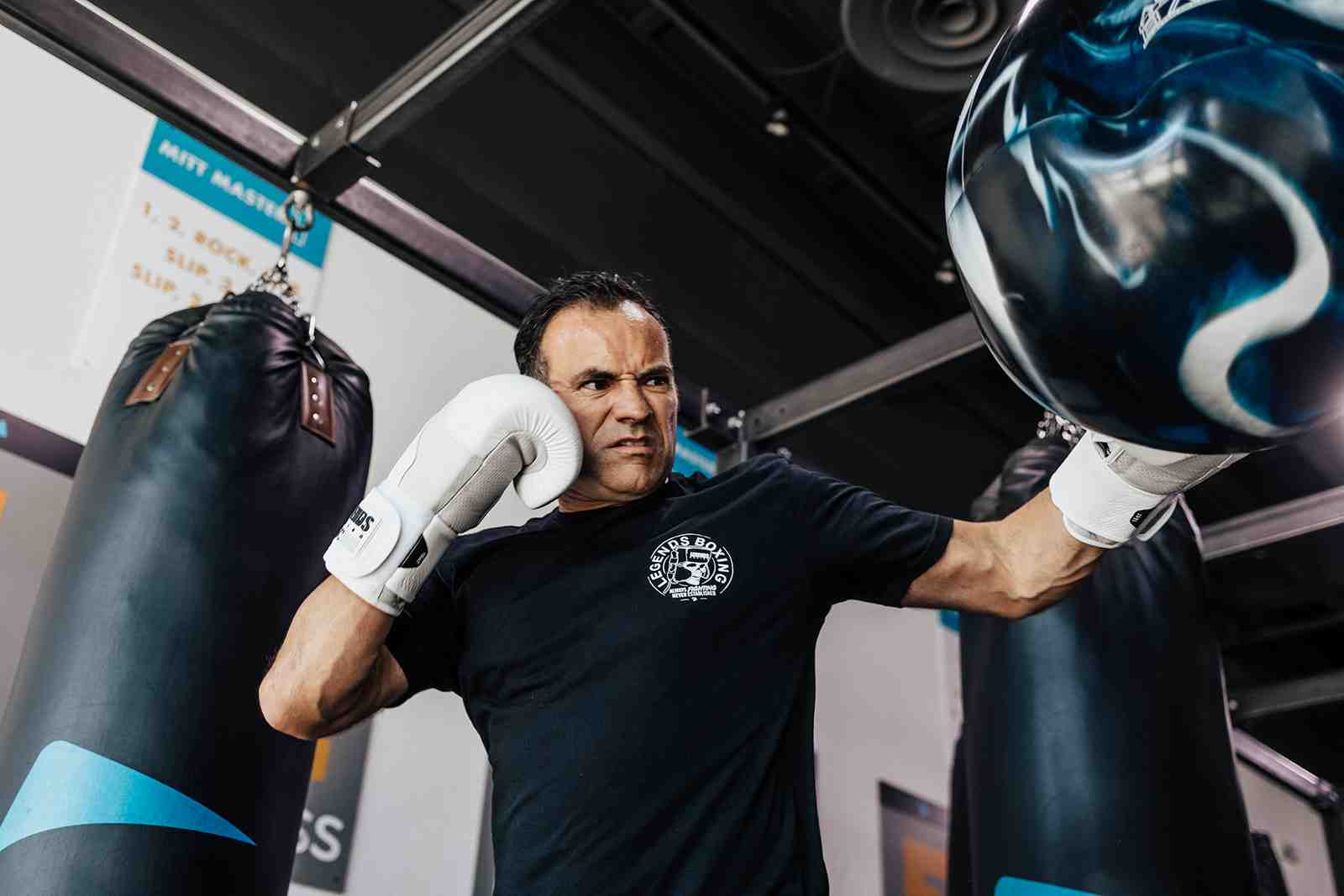 A focused older man wearing a Legends Boxing t-shirt and white boxing gloves lands a sharp punch at a heavy bag, jaw set, eyes locked on target, with a "Mitt Master" combination chart visible on the gym wall behind him — the intensity and technique that comes from consistent practice