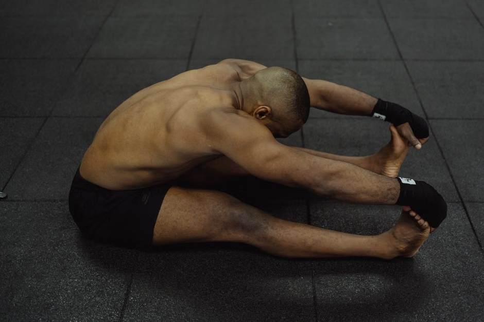A shirtless muscular boxer with black hand wraps sits on a dark rubber gym floor in a deep seated forward fold — legs extended, torso folded over, hands reaching toward his feet — the quiet discipline of a proper post-workout cool-down that prevents injury and accelerates recovery