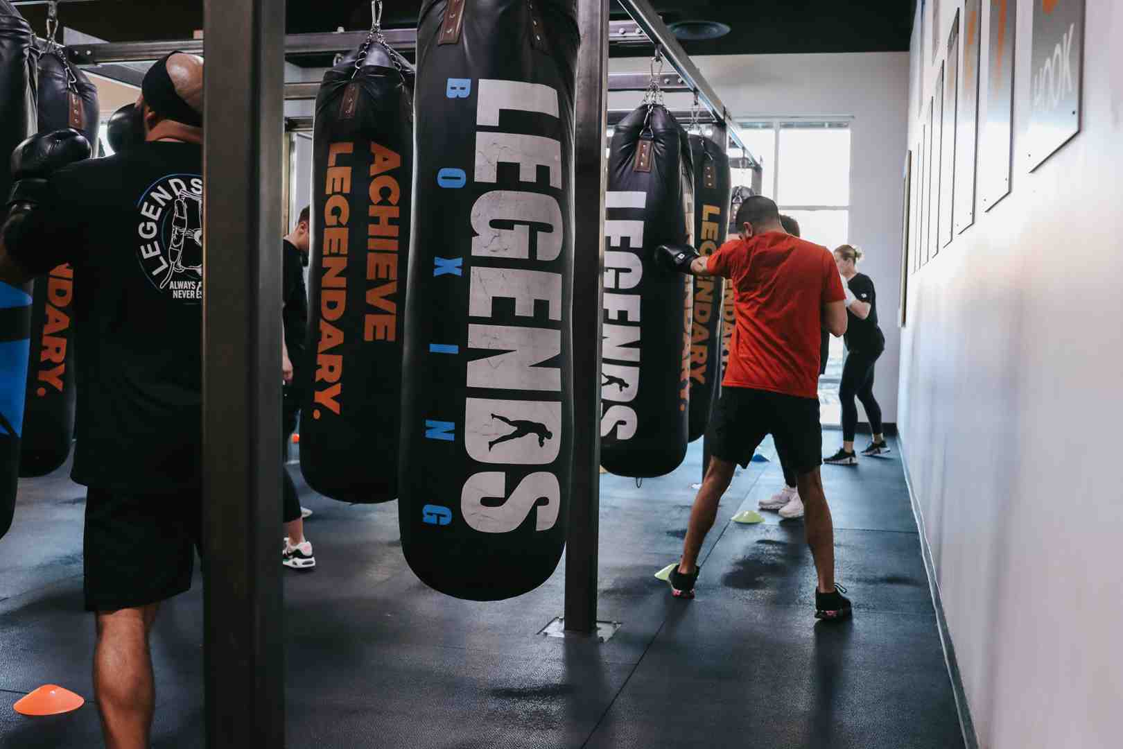 A full row of Legends Boxing heavy bags — "ACHIEVE LEGENDARY" and "LEGENDS BOXING" printed in large letters — hangs from a ceiling rack as multiple members work through their bag rounds in a bright, professionally equipped gym. This is the level of bag setup you're aiming to replicate at home