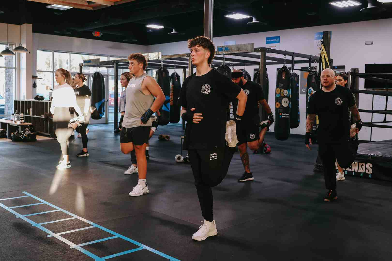 Legends Boxing class students doing warm-up knee-raise lunges in a row across the gym floor, heavy bags and boxing ring visible in the background — showing the full-class warm-up format that prepares your body for the intense work ahead