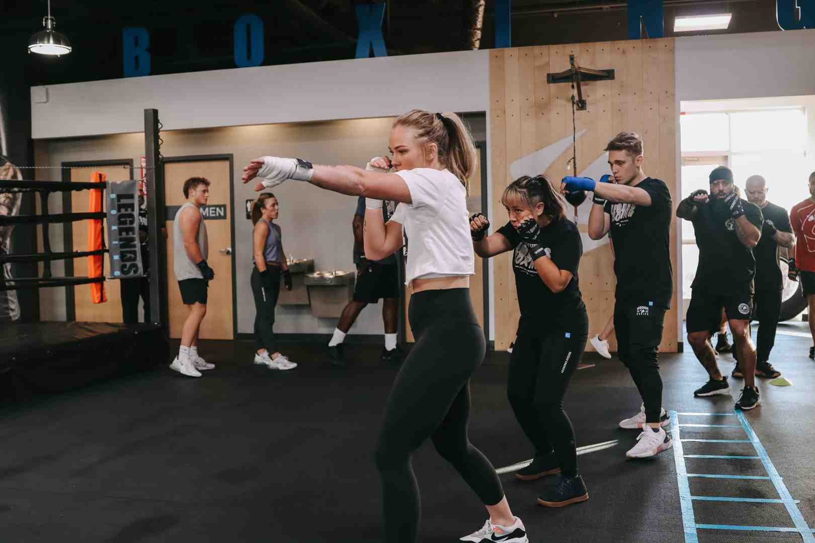 Legends Boxing group class with woman in white top leading the room throwing a cross, multiple students in proper stance visible behind her, "BOXING" sign on the wall — showing the high-energy, coach-led class environment where basics are drilled into muscle memory