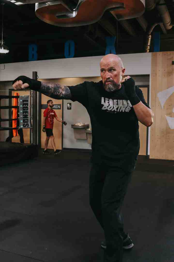 Bald Legends Boxing instructor in a "Legends Boxing" shirt throwing an extended cross with full hip rotation inside the Legends Boxing gym — demonstrating the ground-up power transfer technique with proper form