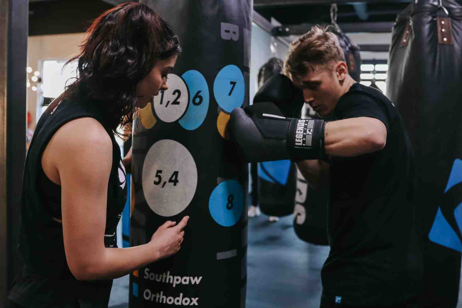Legends Boxing member throwing a punch at a numbered target heavy bag (showing target zones labeled 1,2 / 6 / 7 / 5,4 / 8 for Southpaw and Orthodox) while an instructor watches form — showing how the numbered punch system translates directly to bag work training