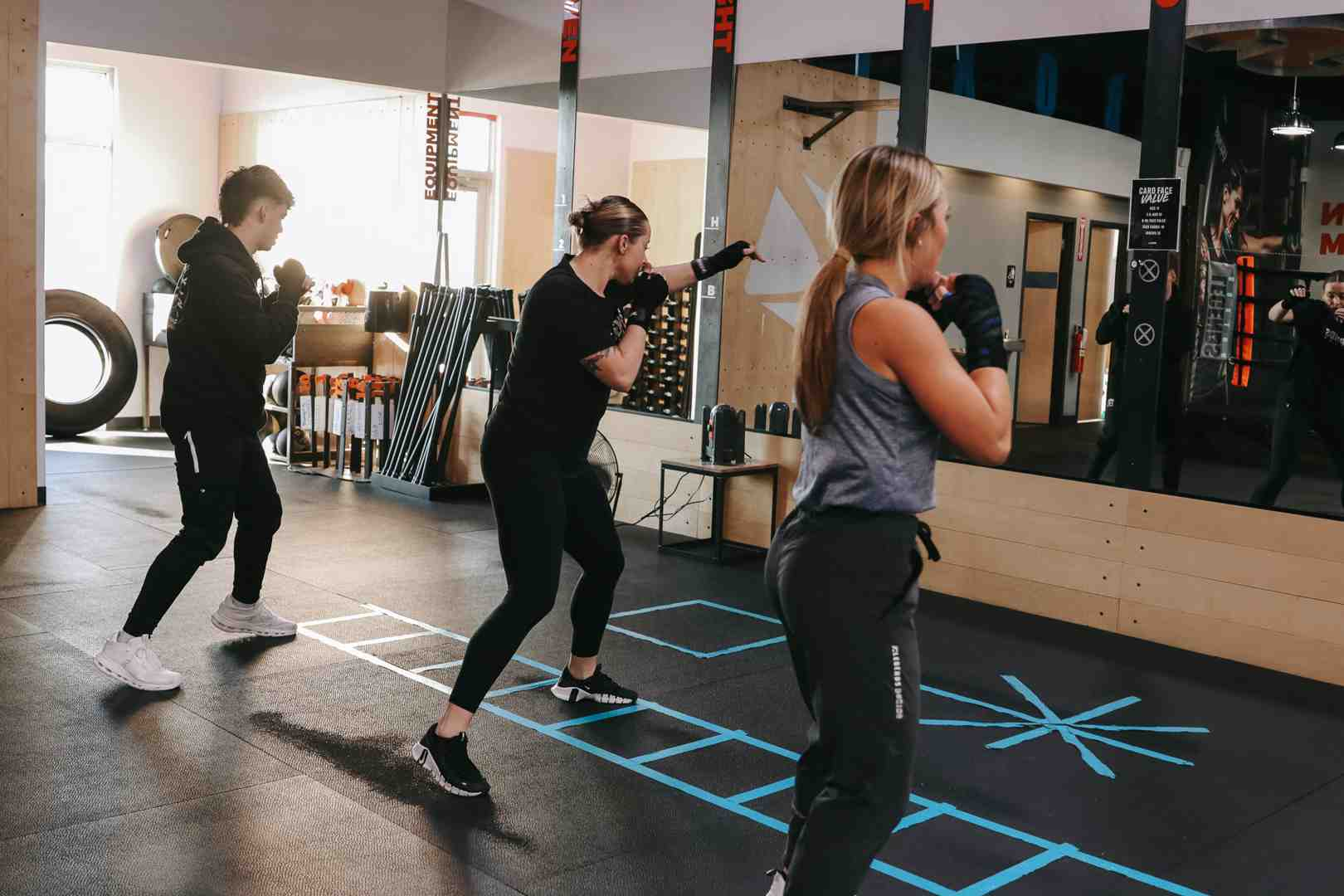 Three students doing boxing footwork drills in a gym with blue tape squares on the floor marking movement patterns, large mirrors reflecting the class — showing the footwork coordination that transforms boxing from arm-only punching into full-body athleticism
