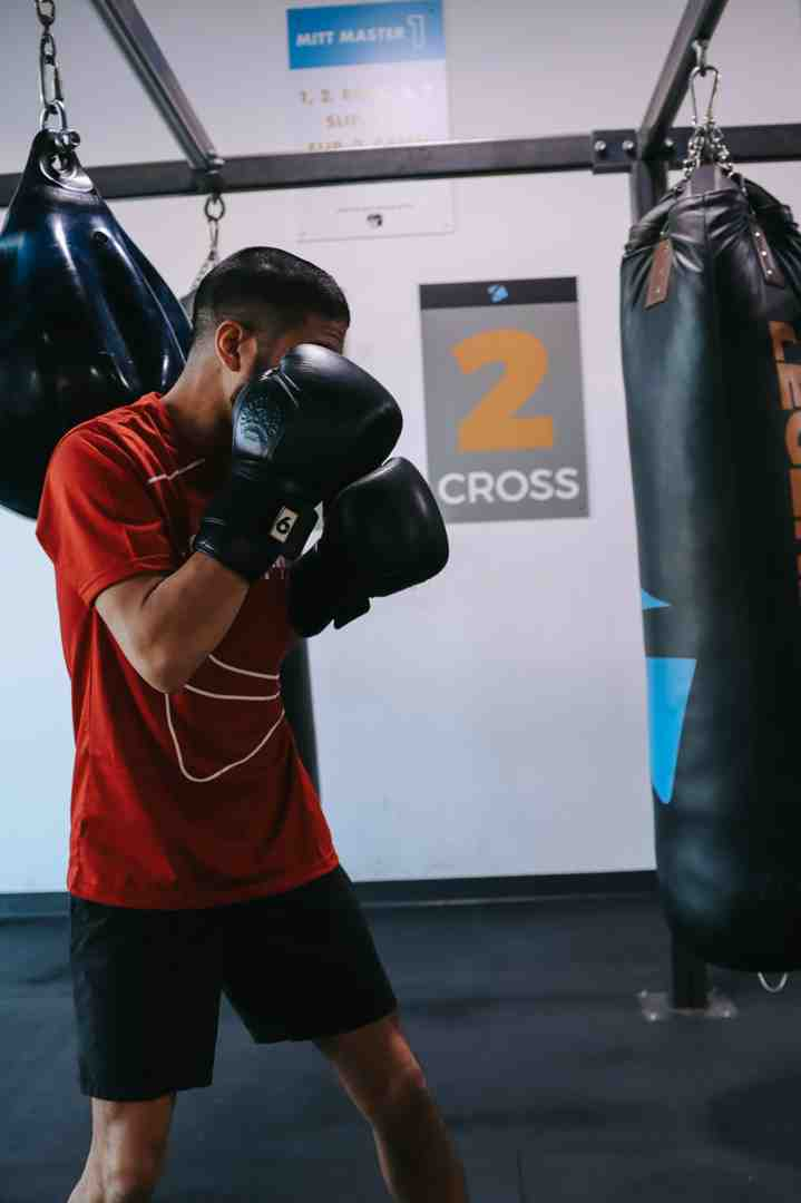A young man in a red t-shirt and black shorts stands in a confident boxing guard at a Legends Boxing gym, both black gloves raised to cheek height in a tight guard, the wall sign directly behind him reading "2 — CROSS" in bold orange, heavy bags hanging on either side — a perfect visual anchor for what proper guard position looks like