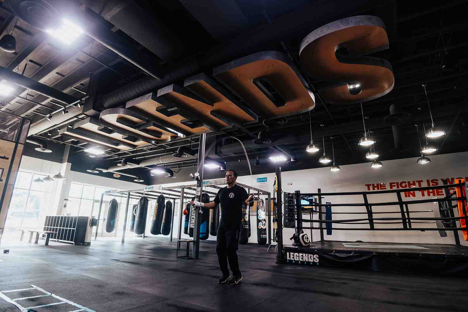 Stunning wide-angle interior shot of a Legends Boxing facility — enormous "LEGENDS" letters hang from a black industrial ceiling above a spacious training floor, boxing ring on the right, rows of heavy bags on the left, a trainer jumping rope in the center, "THE FIGHT IS WON IN THE..." painted on the far wall — showing the professional-grade gym environment Legends Boxing Houston members train in
