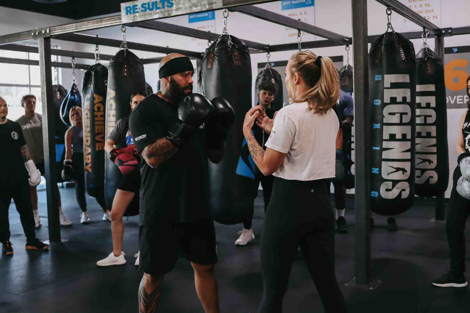 Legends Boxing instructor wearing a headband and boxing gloves working with a female student on technique, other class members watching in the background, "ACHIEVE LEGENDARY" branded heavy bags and rows of Legends-branded bags visible — showing the focused one-on-one coaching style that makes Legends Boxing classes effective for all skill levels