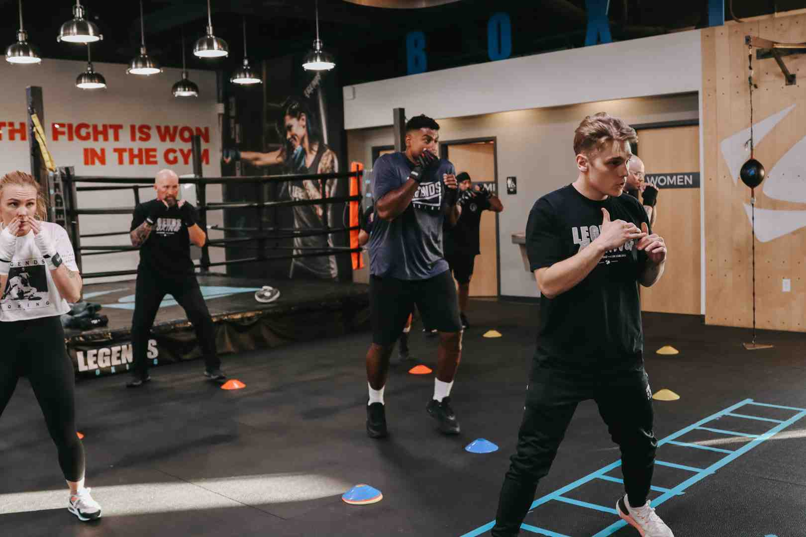 Energetic Legends Boxing class in full swing — 'THE FIGHT IS WON IN THE GYM' mural on the wall, members in boxing stance on agility floor, boxing ring and 'LEGENDS' branding in background — Salt Lake City