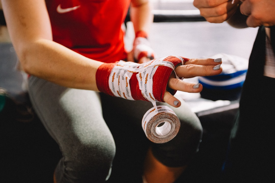 Close-up of a coach helping a female athlete wrap her hands in red and white hand wraps before a boxing class — protective hand wrapping technique, gym ring in the background