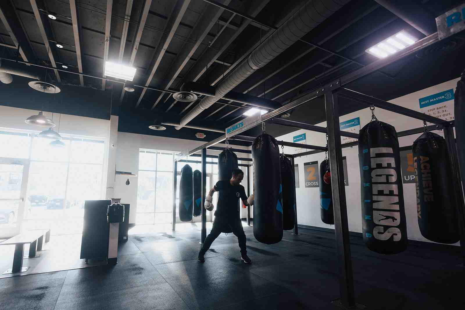 Wide-angle interior of a Legends Boxing gym — solo boxer working heavy bags labeled 'LEGENDS' and 'ACHIEVE', natural light from large windows, 'CROSS' and 'MITT MASTER' instruction signs on the walls — Legends Boxing Utah