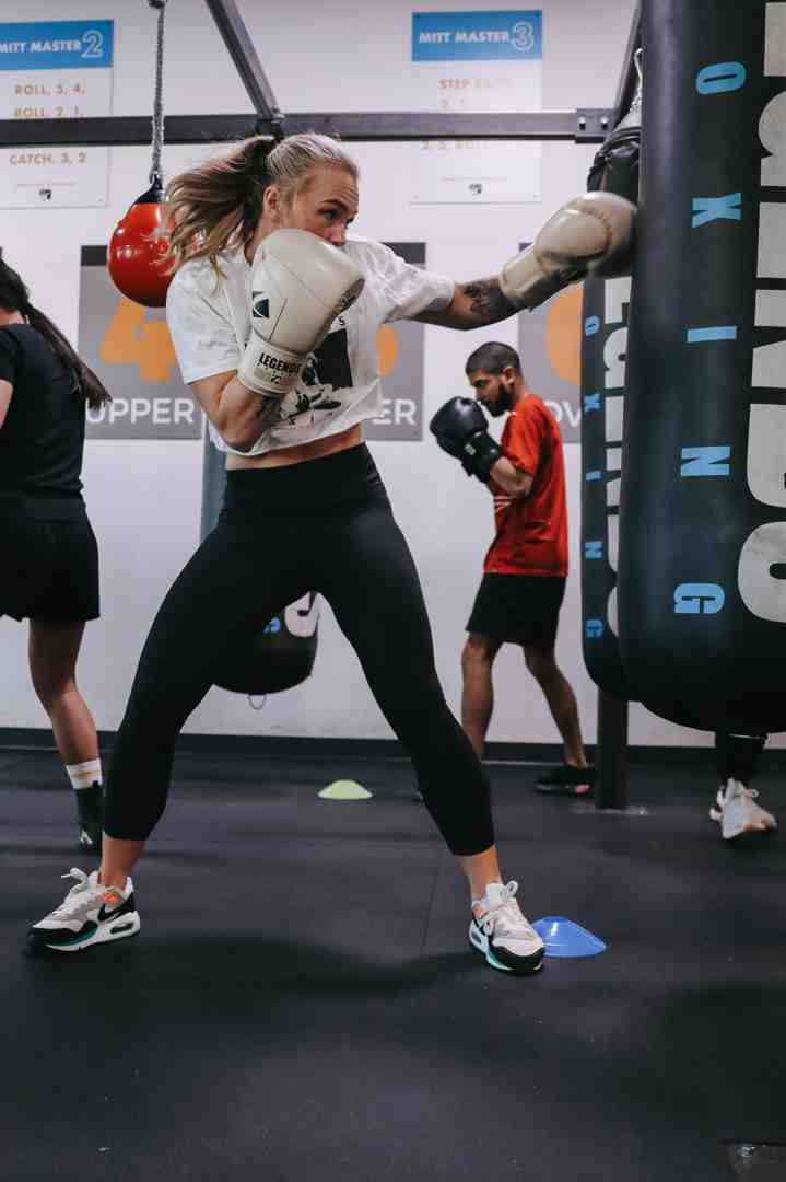 Woman wearing Legends Boxing gloves throwing a cross at a heavy bag — 'UPPER' and 'MITT MASTER' instruction signs on the wall, other boxers training in the background — Legends Boxing Utah class