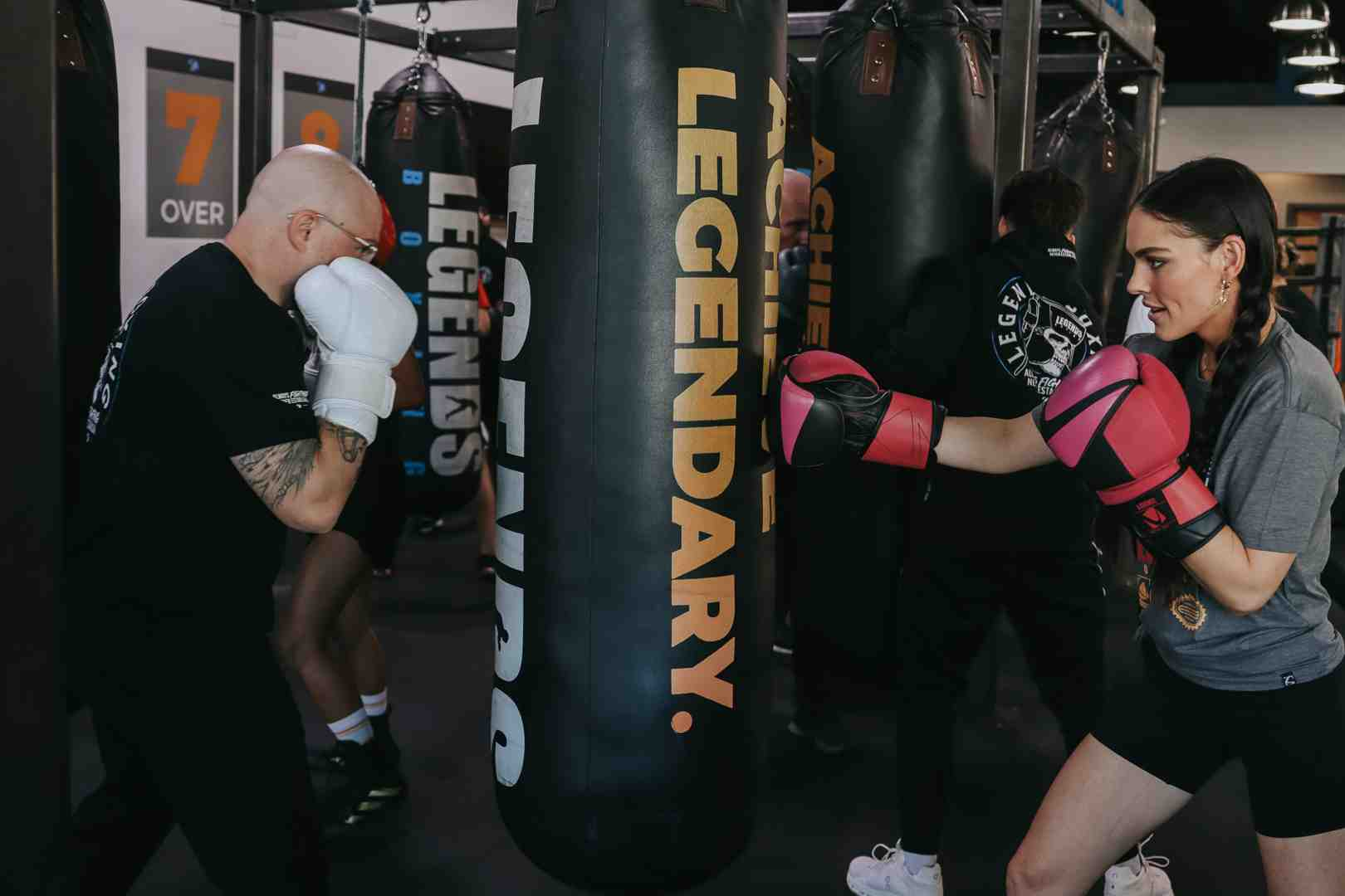 Tattooed bald man in white boxing gloves and woman in pink boxing gloves both punching "Achieve Legendary" branded Legends Boxing heavy bags side by side in the gym — showing the high-energy, side-by-side bag work format that characterizes a Legends Boxing class session