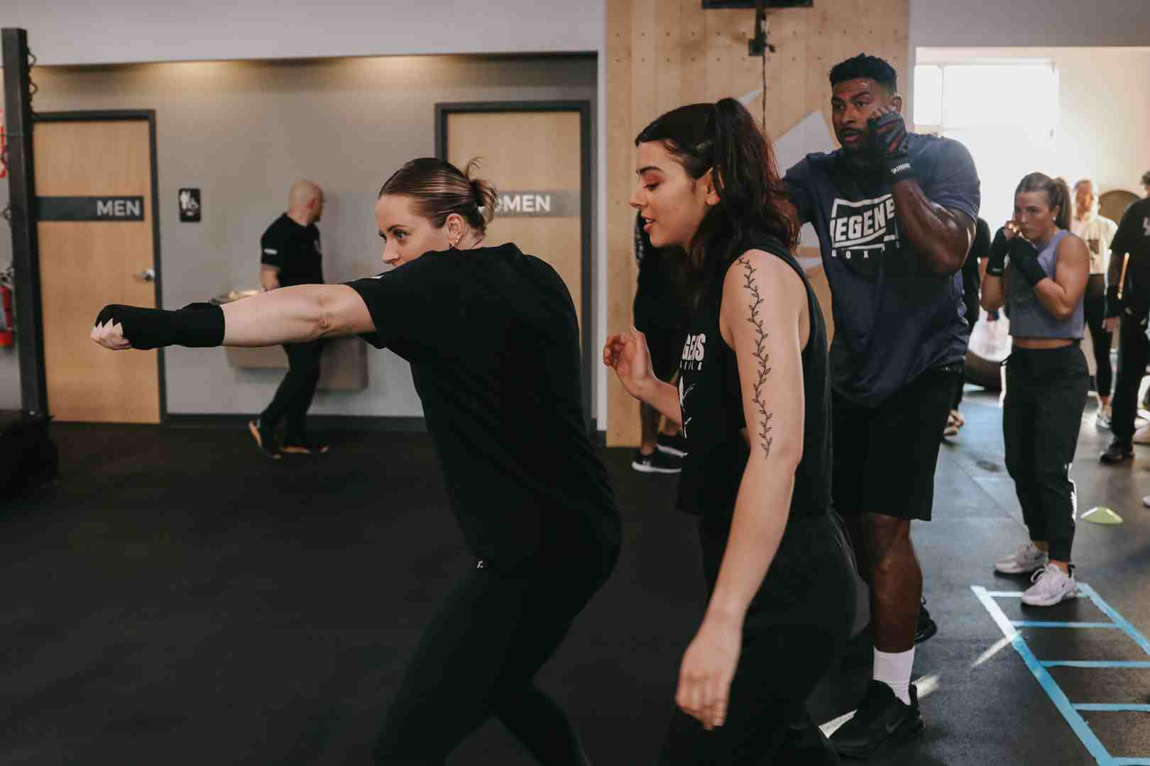 Two Legends Boxing women students throwing punches in class with a male Legends Boxing instructor watching their form from behind — showing the attentive coaching and side-by-side partner drilling that accelerates technique development