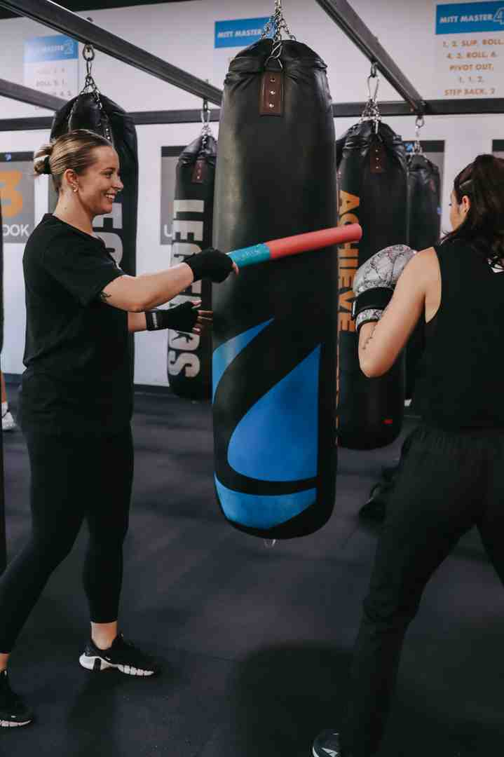 Two women at Legends Boxing — one smiling in black holding a red foam stick against the blue Legends Boxing signature bag while the other hits the ACHIEVE bag in the background — Mitt Master combination drills visible on the wall signs — Legends Boxing