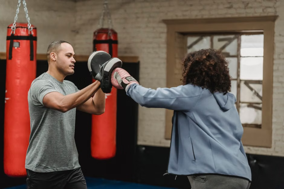 Coach holding grey focus mitts while a woman in pink boxing gloves and a blue hoodie throws a jab — mitt work drill in a boxing gym with red heavy bags and white brick walls visible — Legends Boxing