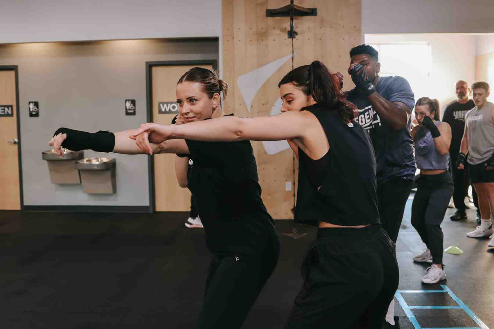 Two women in black workout gear practice together in a Legends Boxing class, both extending their arms in a synchronized punch-drill with perfect mirrored form, while a Legends Boxing coach in a dark t-shirt guides the session behind them — the kind of partner training that builds defensive reflexes and counter-punch timing