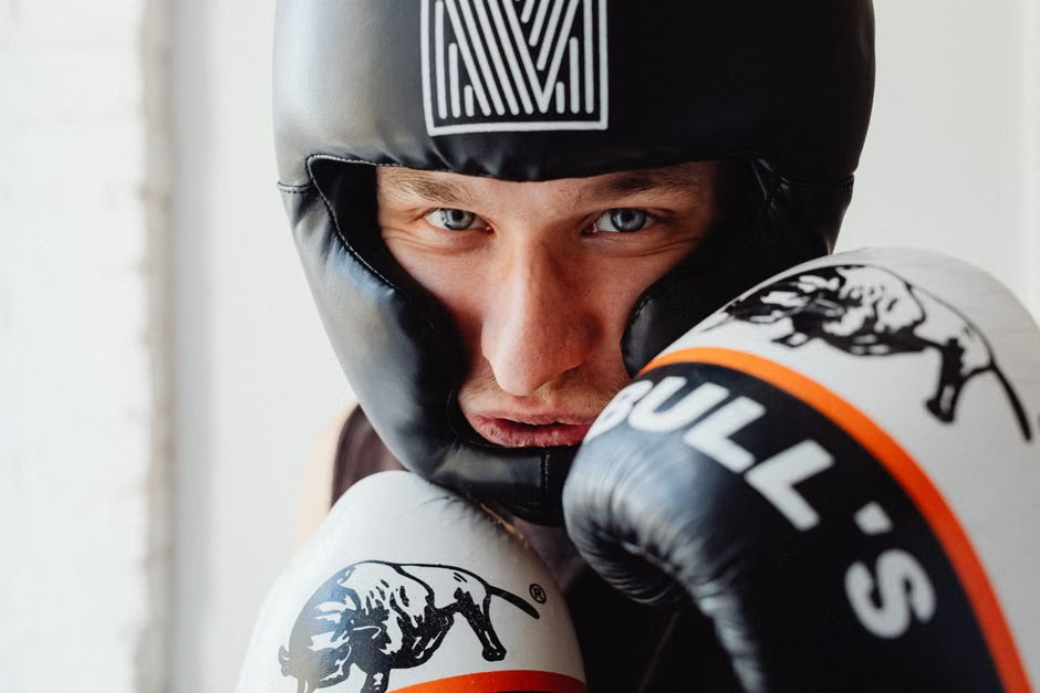 A boxer wearing sleek black matte headgear and Bull's boxing gloves stares directly at the camera with intense blue eyes, gloves raised in a tight high guard position — the protective gear and discipline that good defense is built on