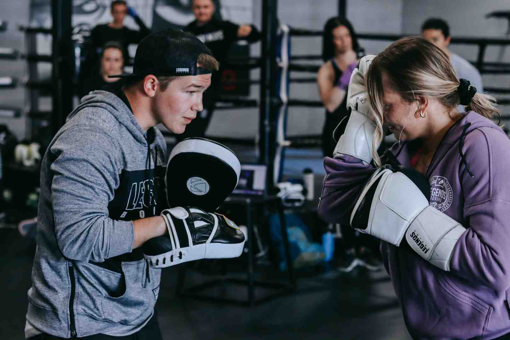 A Legends Boxing trainer in a gray hoodie and cap holds large focus mitts as a student in a purple Legends Boxing hoodie uses white gloves to practice a defensive guard block, with athletes watching in the background of a dark, well-equipped boxing gym