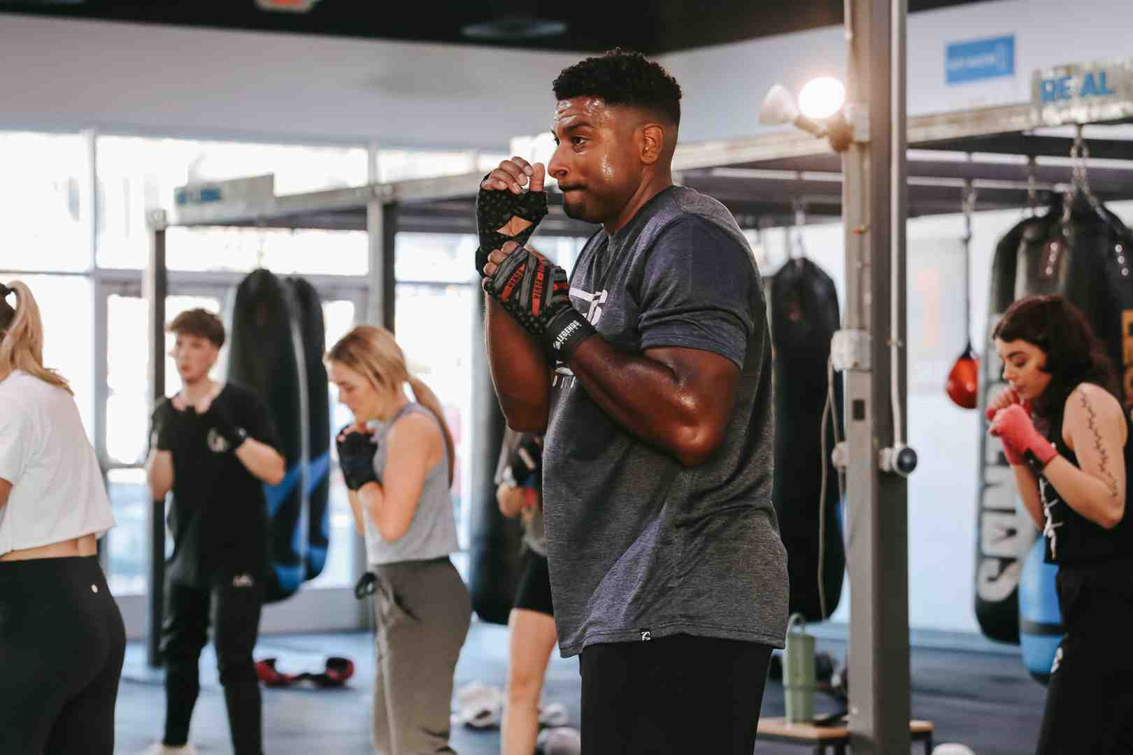 Diverse group of Legends Boxing members in guard position during a class — man center-frame in training gloves, multiple members and heavy bags visible throughout the bright gym — boxing class community — Legends Boxing