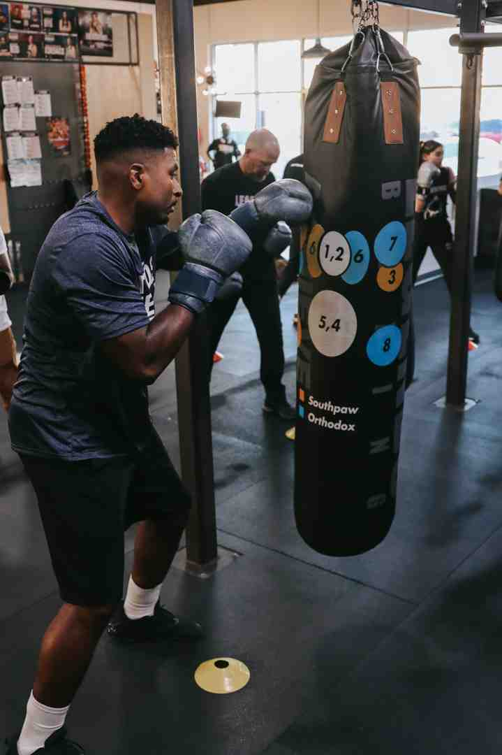 Member throwing a punch at a numbered target heavy bag with proper technique — Legends Boxing coach and other members training in background — bag work training gloves in action — Legends Boxing
