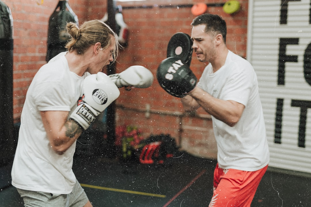 Two men in white t-shirts doing intense pad work, sweat flying mid-air as one drives a right hand into a round pad held by his partner, brick wall and gym signage visible in the background — the kind of focused one-on-one coaching Savarese Fight Fit is known for