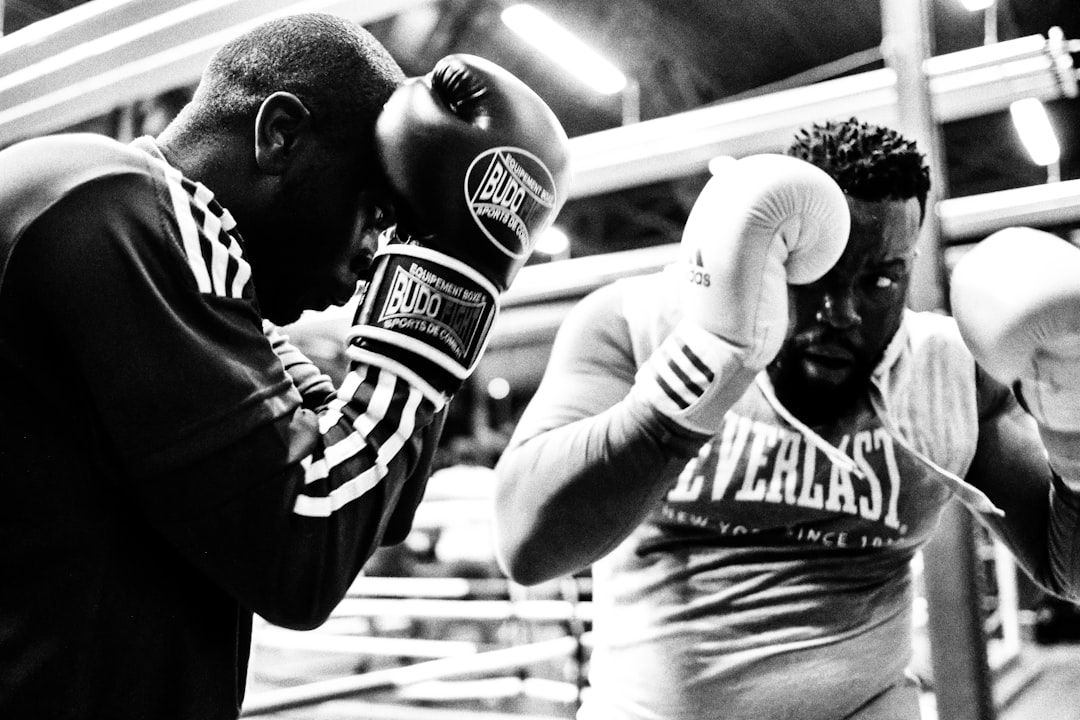Black and white photograph of two heavyweight fighters sparring in a boxing ring — one in Budo Fight gear landing against the other in an Everlast top, both in tight guard, gym lights glowing overhead — the raw intensity of traditional boxing training that made legendary gyms like Hank's what they are