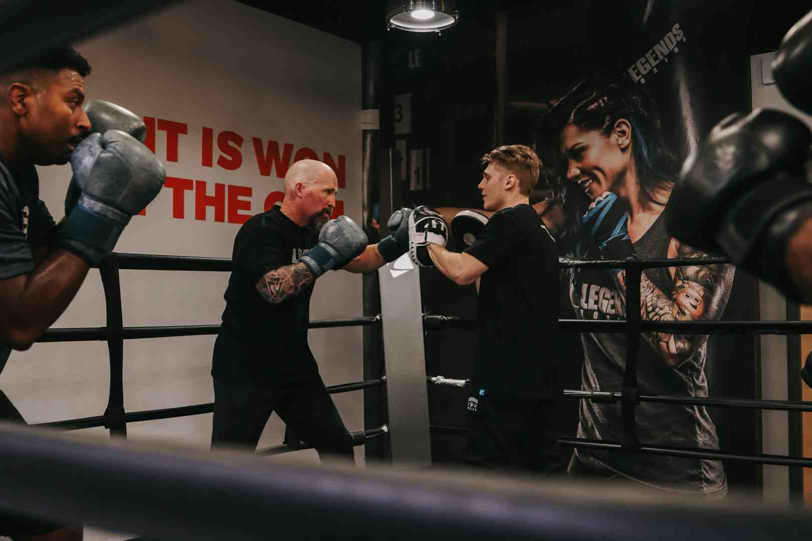 Inside the Legends Boxing ring — bald tattooed coach sparring with a young male member, 'IT IS WON IN THE GYM' mural and Legends Boxing athlete poster visible on the wall behind them — Salt Lake City