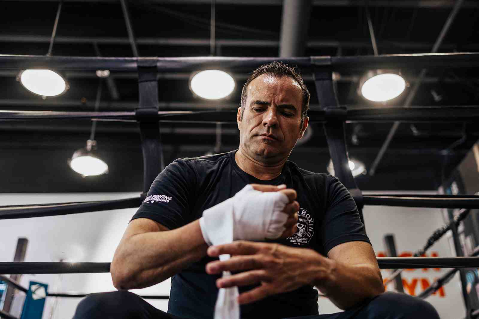 Male Legends Boxing member sitting near the boxing ring, carefully wrapping hands in white hand wraps, 'Legends Boxing — Always Fighting Never Established' logo on shirt — Utah County gym