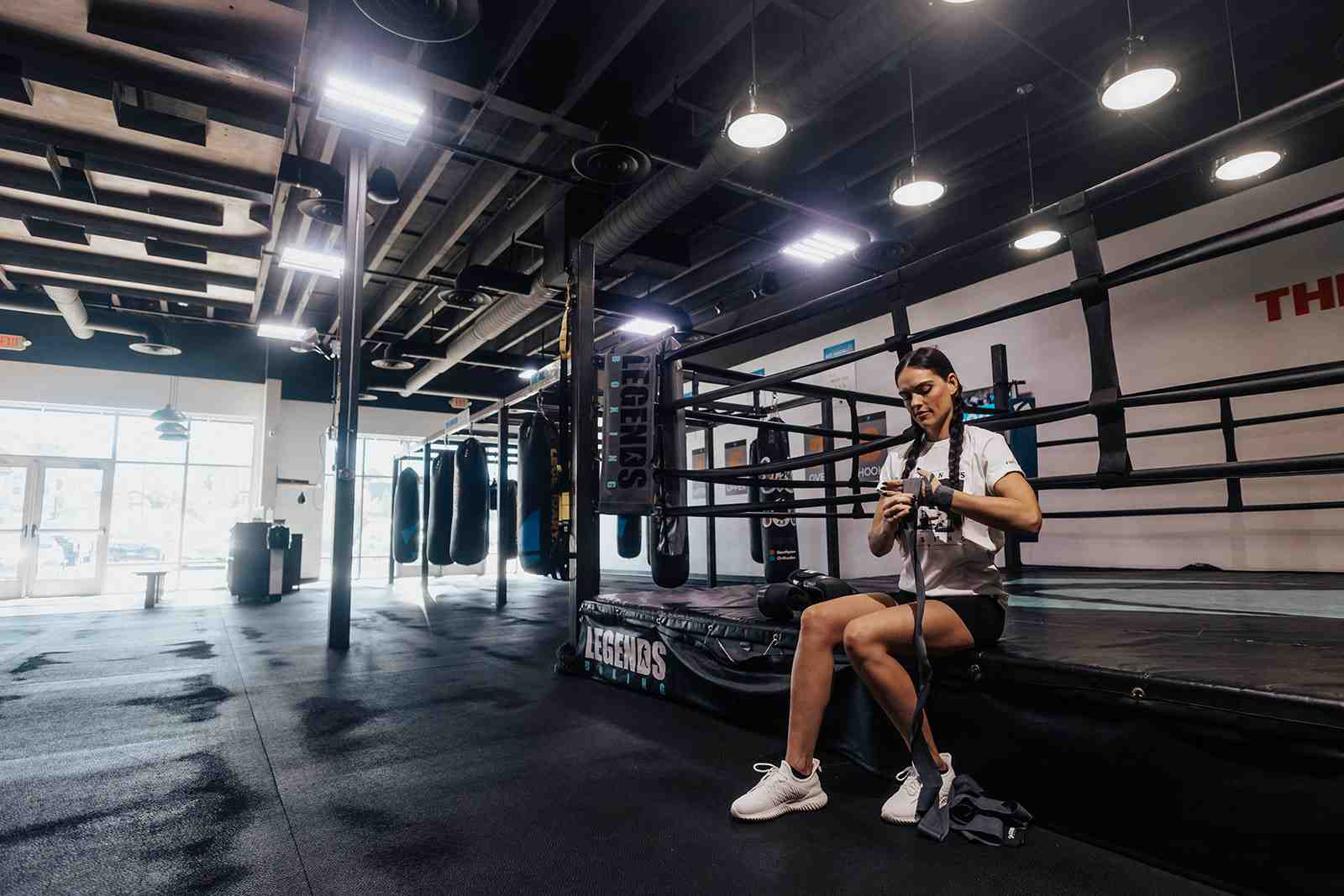 Woman sitting on the edge of a Legends Boxing ring carefully wrapping her hands, heavy bags visible in background, clean modern Utah gym space