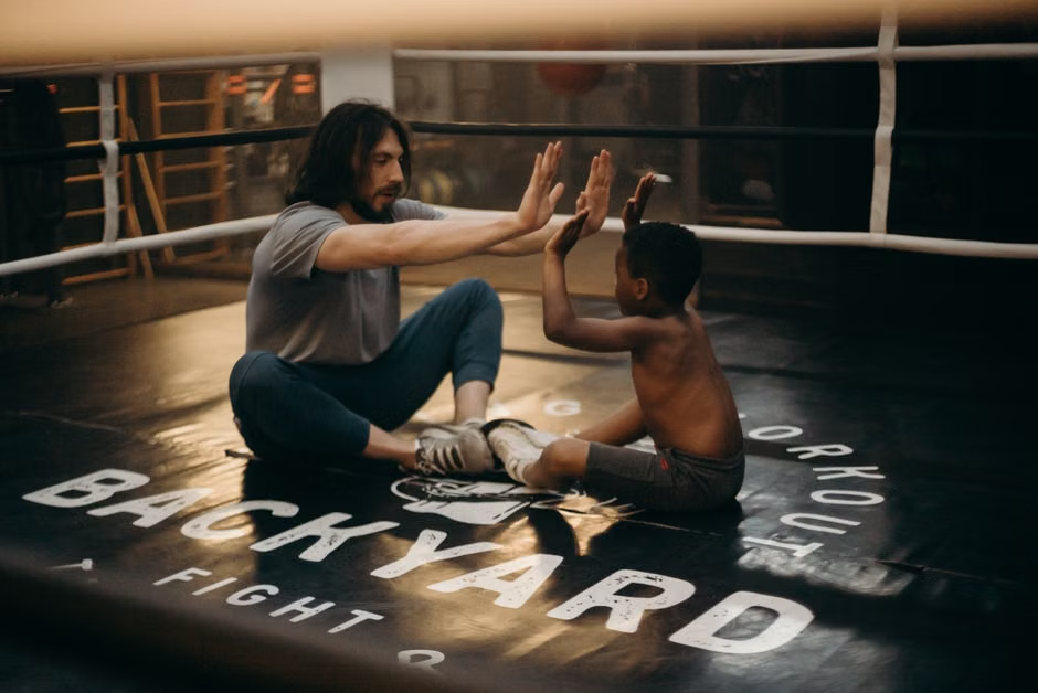 A long-haired adult coach and a young child sit cross-legged face-to-face in the center of a boxing ring, both with hands raised and palms pressed together in a high-five drill — a warm, playful training moment that captures the mentorship and trust at the heart of youth boxing programs