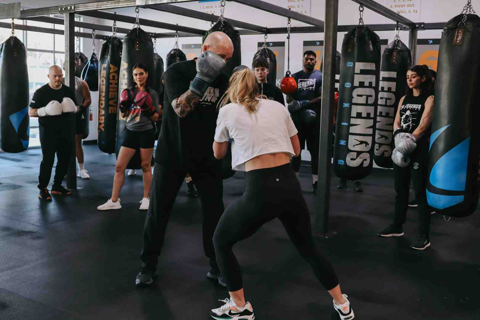 A bald tattooed Legends Boxing coach demonstrates close-range technique with a female member in a white top, glove-to-glove contact in a coaching drill, while other members in black attire observe from stations around the gym — "ACHIEVE LEGENDARY" and "LEGENDS" bags visible throughout the well-lit space