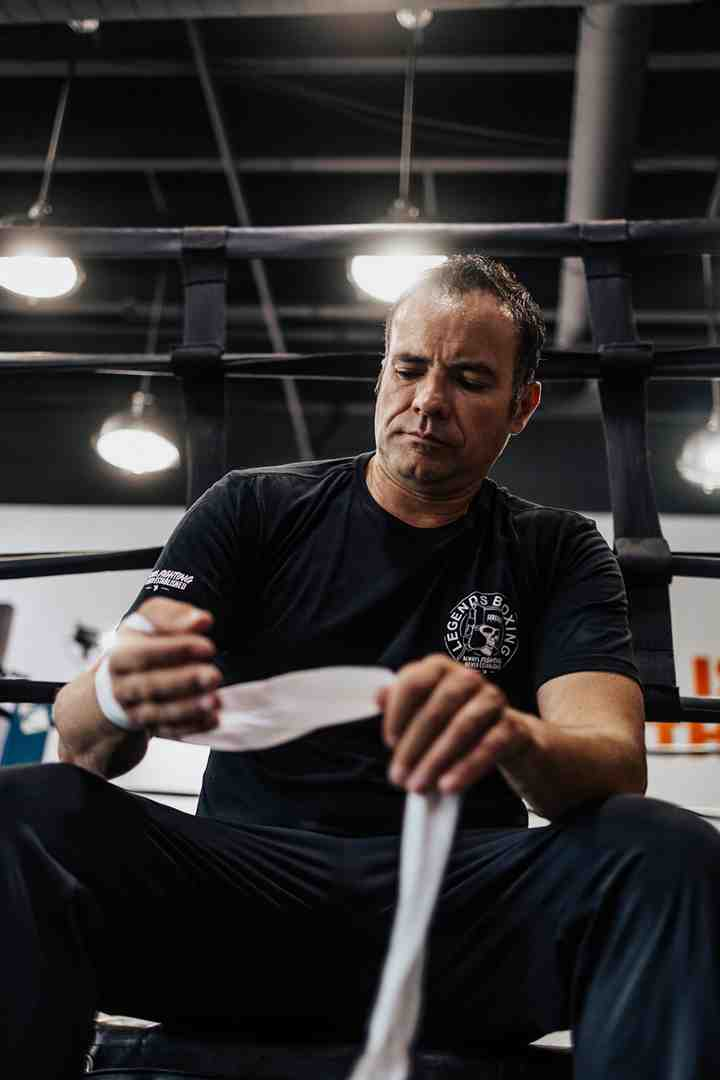 A man in a Legends Boxing t-shirt sits inside a boxing ring, head bowed in concentration as he carefully wraps white hand wraps around his hands, the ring's black ropes framing him and bright gym pendant lights glowing overhead — the pre-session ritual that protects your 27 small hand bones every time you train