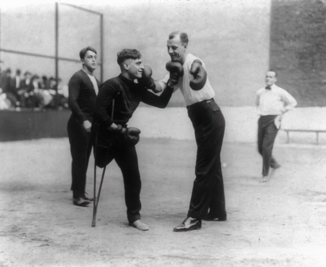 Early 20th-century black-and-white photograph of two young men sparring outdoors with boxing gloves — one using a cane for balance, the other in guard — two onlookers watching, spectators visible in background — a rare glimpse of boxing history in practice — Legends Boxing