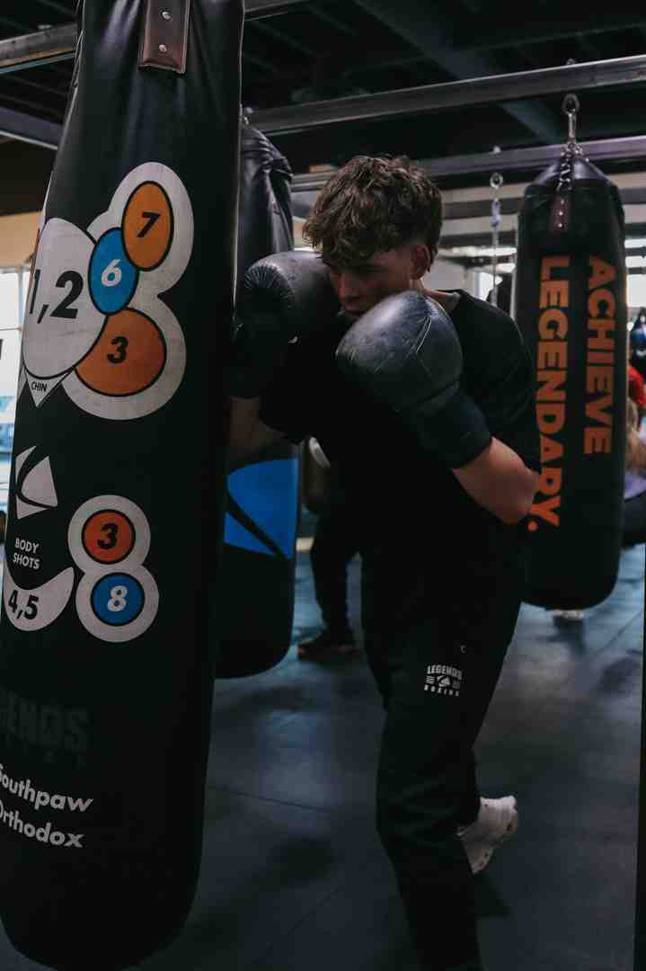 Young man with curly hair throwing a combination at a numbered Legends Boxing heavy bag — colored target circles showing 1,2 / 6 / 3 / 7 punch zones, body shot targets (4,5 / 3 / 8) below — "ACHIEVE LEGENDARY" heavy bag visible on right — showing proper close-range bag work technique for beginners