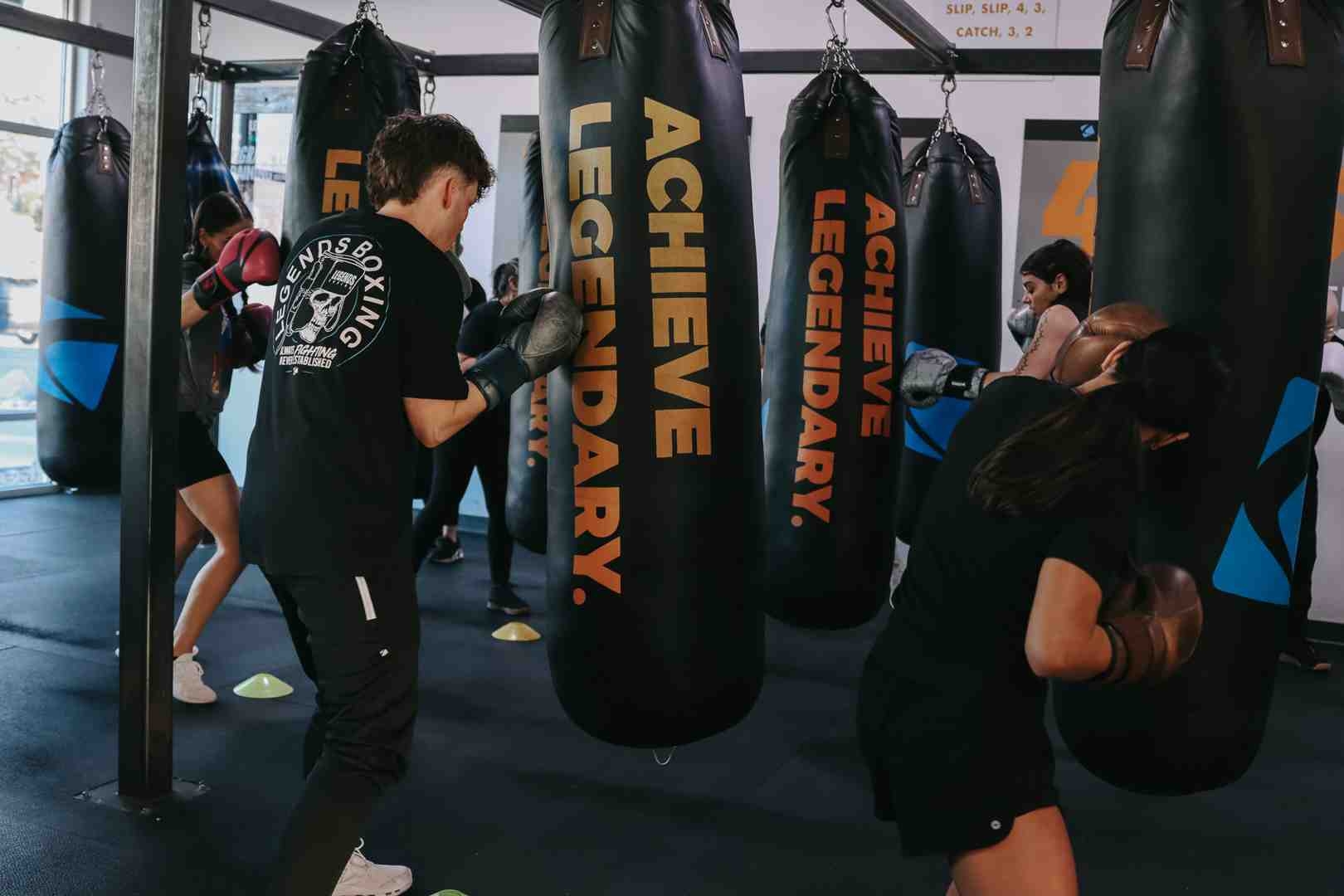 Legends Boxing group class hitting 'ACHIEVE LEGENDARY.' heavy bags — coach in foreground, mirror wall behind, 'SLIP, SLIP 4,3 / CATCH 3, 2' drill card visible on wall — energetic group environment