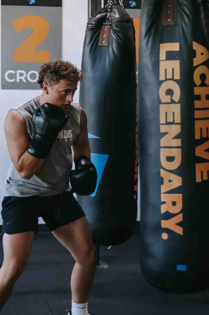 Young man in Legends Boxing gear throwing a cross at an 'ACHIEVE LEGENDARY.' heavy bag — '2 CROSS' sign visible on the wall in the background — Legends Boxing gym