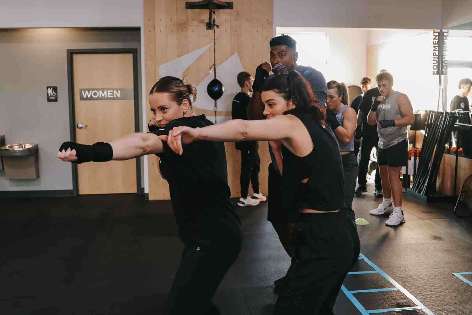 Legends Boxing class in session — two women throw simultaneous punches in matching stance while coach in gray shirt guides them from behind — several other members training in the bright, welcoming gym in the background — boxing lessons near me at Legends Boxing