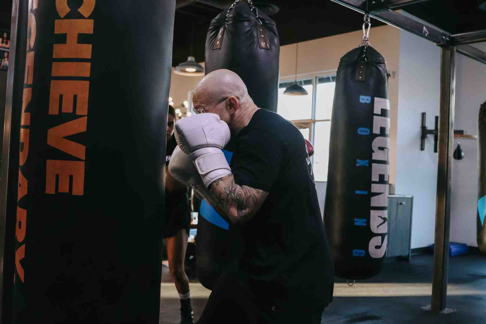 Bald boxer in black Legends Boxing shirt working close range on a heavy bag at a Legends Boxing gym — "ACHIEVE LEGENDARY" branding on the bag, mirror reflection and additional heavy bags visible — boxing moves to learn: defensive positioning and bag work — Legends Boxing