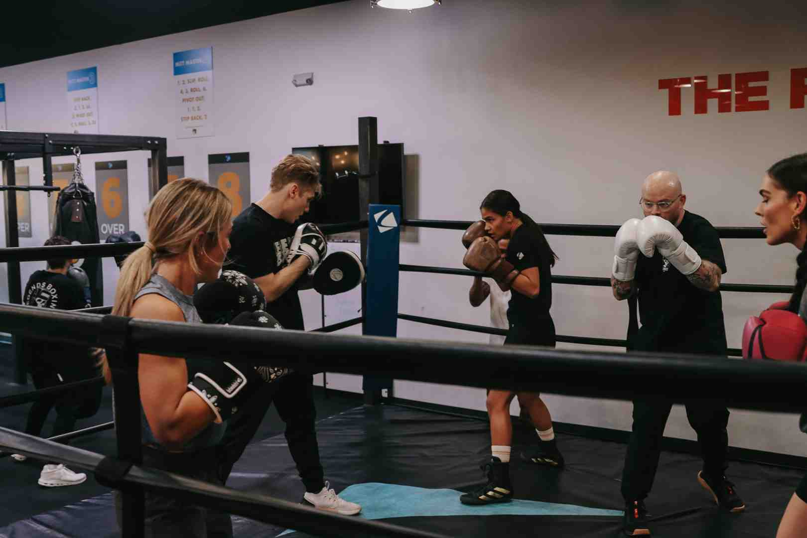 Multiple people doing mitt work with coaches inside a boxing ring at a Legends Boxing gym — overhead number displays visible on the wall behind them, everyone wearing gloves and working in pairs — capturing the energy of a beginner-friendly boxing class