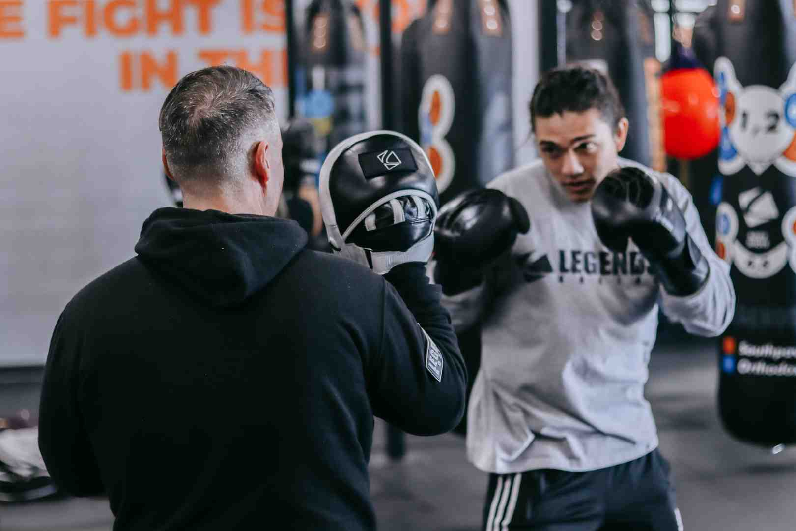 Trainer holding a focus mitt while a student in a Legends Boxing sweatshirt throws a punch — "THE FIGHT IS IN THE..." wall mural visible in background — heavy bags and a double-end ball hanging in the gym — showing one-on-one pad work for beginners learning basic boxing combinations