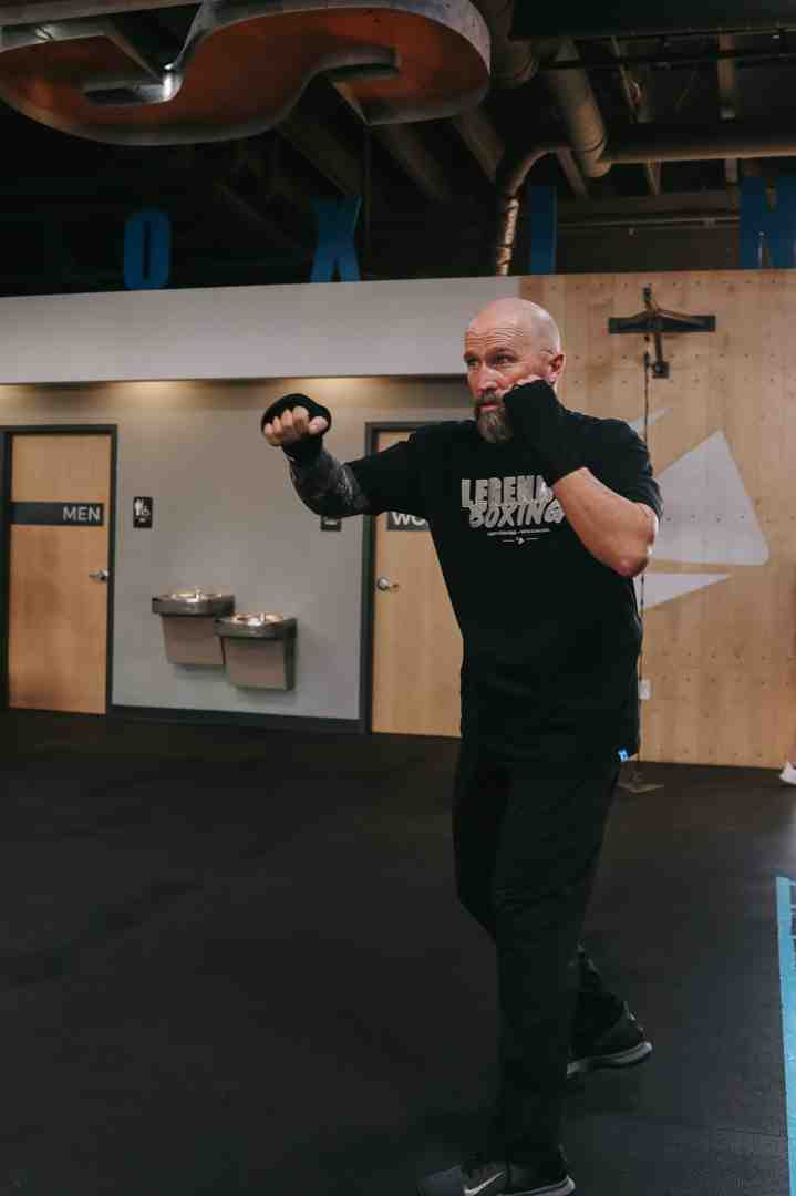Bald man in a Legends Boxing t-shirt demonstrating a jab in shadowboxing stance inside a Legends Boxing gym — "BOXING" lettering and "OX" wall art visible behind him — showing the proper extension and guard position for basic boxing moves for beginners