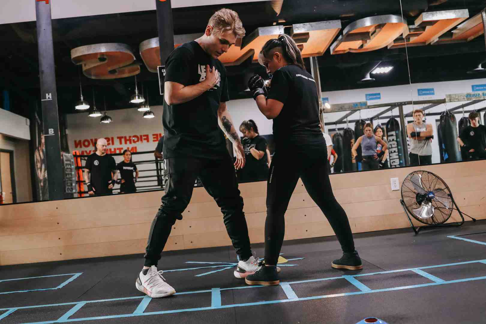 Legends Boxing coach in all-black guiding a female student through footwork drills on a marked mat — mirror wall reflecting the full class in session behind them — hands-on technique correction at Legends Boxing — boxing moves coaching