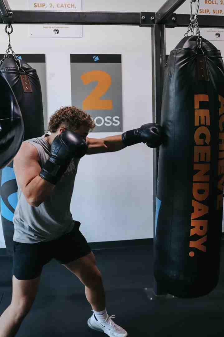 Curly-haired man in grey Legends tank top throwing a powerful cross at an "ACHIEVE LEGENDARY." heavy bag — "2 CROSS" sign on wall behind — full body rotation, weight transferred forward, classic cross mechanics — boxing moves at Legends Boxing