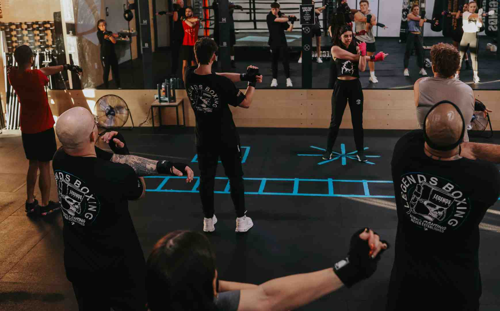Legends Boxing class in session — members in Legends Boxing shirts shadowboxing on a teal-marked floor with mirrors showing the full room, a coach leading from the front