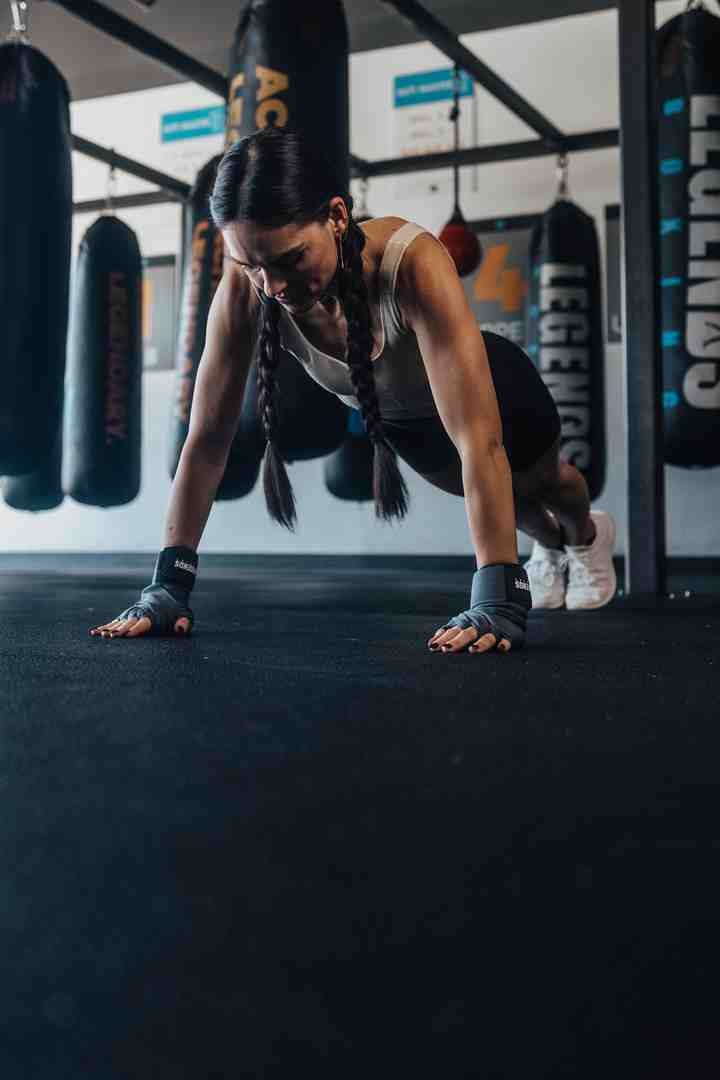 Female boxer with braids doing a push-up on the Legends Boxing gym floor with heavy bags hanging in the background — demonstrating strength training between bag rounds