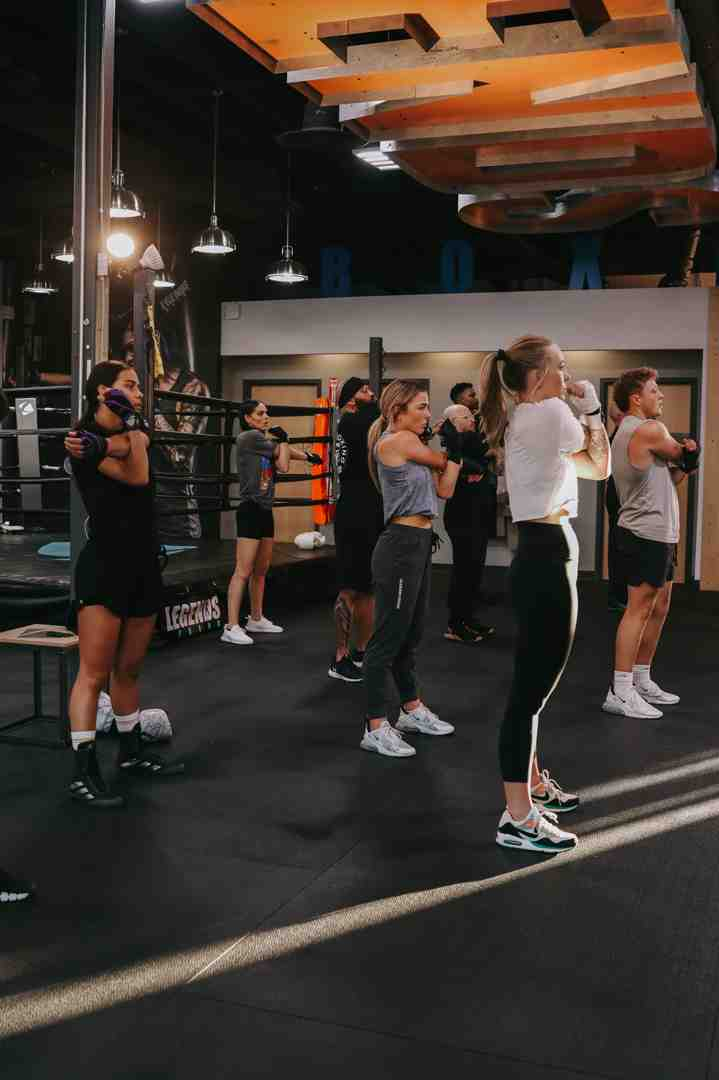 Group boxing class at Legends Boxing — diverse group of men and women in athletic wear standing in boxing stance facing the front, "LEGENDS" visible on equipment, warm-lit gym with orange accent ceiling panels — Draper area boxing fitness