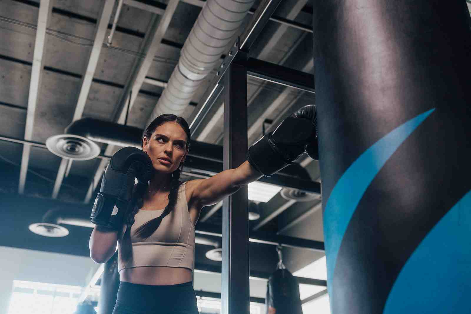 Woman in athletic wear throwing a punch at a large heavy bag — low-angle dramatic shot, industrial gym ceiling, focused determined expression — female boxing training