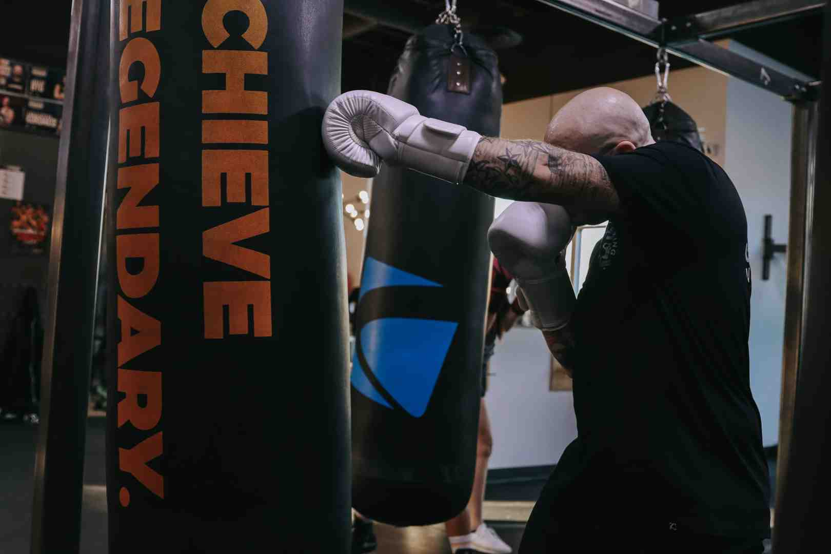 Bald tattooed boxer in black Legends Boxing shirt throwing a left hook into an ACHIEVE LEGENDARY heavy bag — blue Legends-branded bag visible beside him, gym equipment in background — fighting gyms near me: intense bag work delivering real results — Legends Boxing
