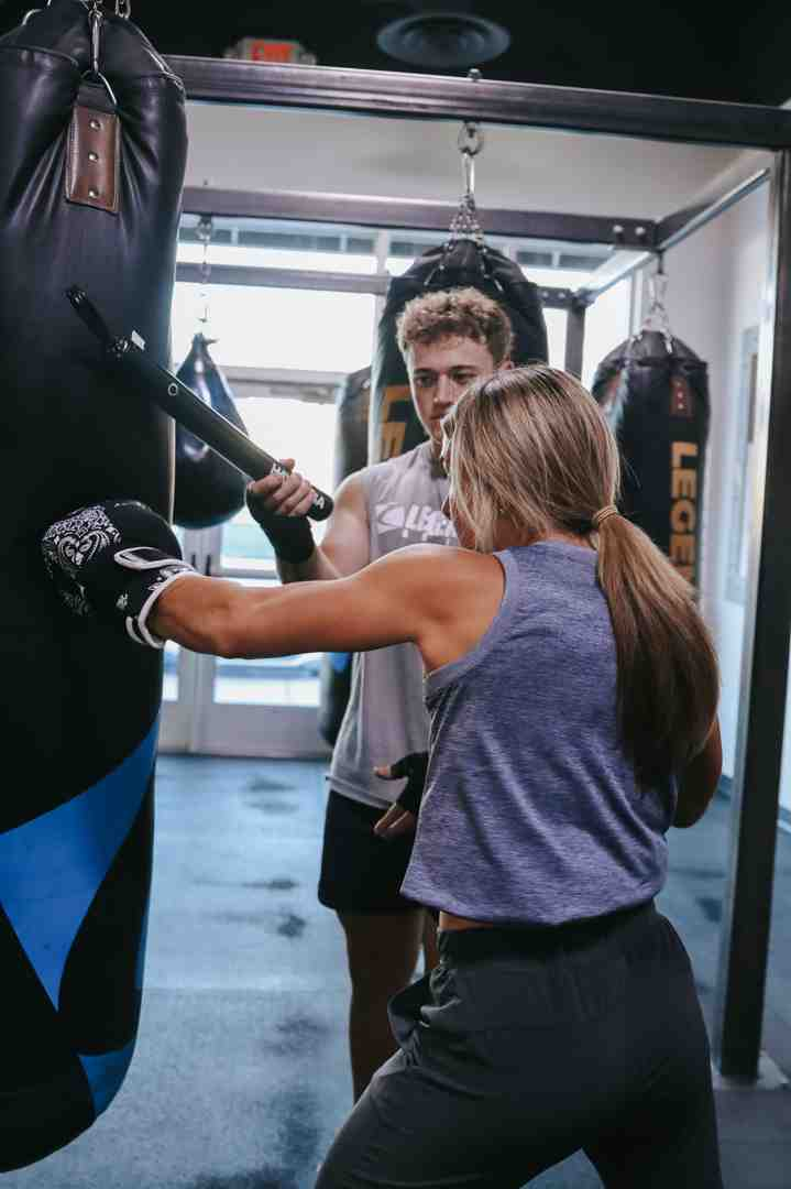 Male Legends Boxing coach in gray shirt holding a pad while female member in purple tank top throws a punch — multiple bags and bright gym windows visible in the background — fighting gyms near me: what great coaching looks like at Legends Boxing