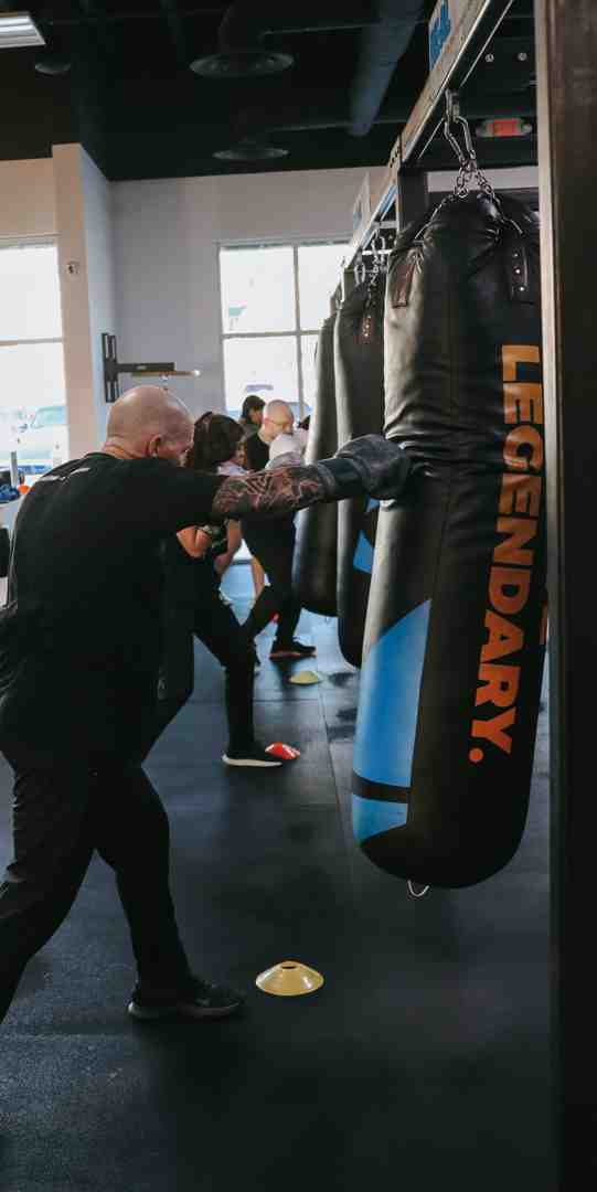 Bald tattooed coach in all-black demonstrating powerful bag work on a LEGENDARY-branded heavy bag at Legends Boxing — multiple members training on bags behind him in a bright, open gym — fighting gyms near me: Legends Boxing class energy — Legends Boxing
